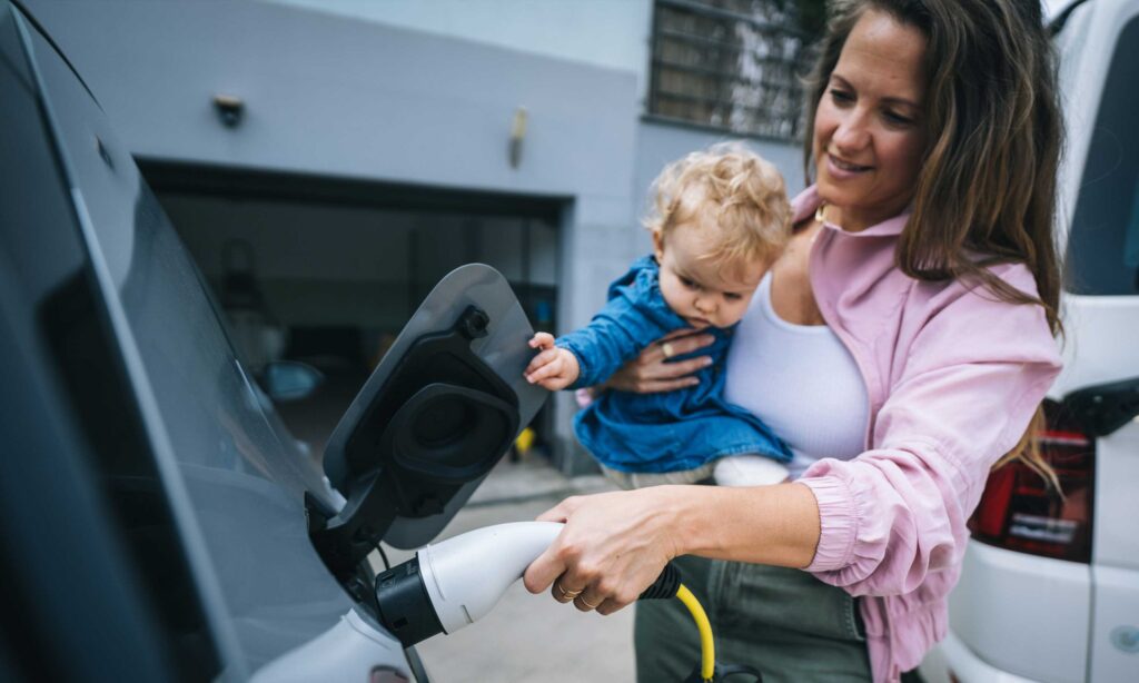 A woman holding a baby and plugging in an electric vehicle